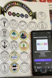 A phone displaying the audiobook, "Melissa" written by Alex Gino next to a partially complete book bingo card.