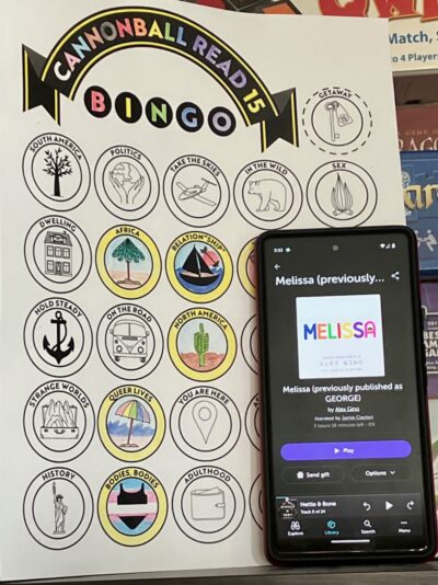 A phone displaying the audiobook, "Melissa" written by Alex Gino next to a partially complete book bingo card.