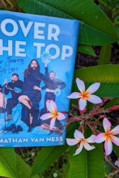 The cover of the book, "Over the Top: A Raw Journey to Self-Love" by Jonathan Van Ness is held up next to cluster of plumeria flowers.