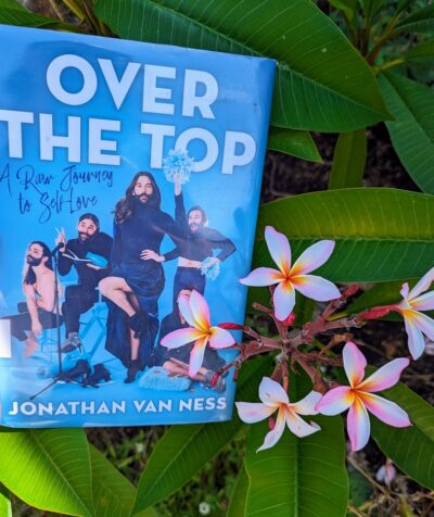 The cover of the book, "Over the Top: A Raw Journey to Self-Love" by Jonathan Van Ness is held up next to cluster of plumeria flowers.
