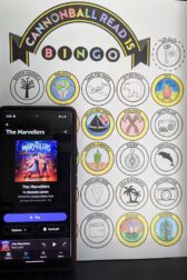 A phone displaying the audio book, "The Marvellers" by Dhonielle Clayton is standing next to a partially complete book bingo card.