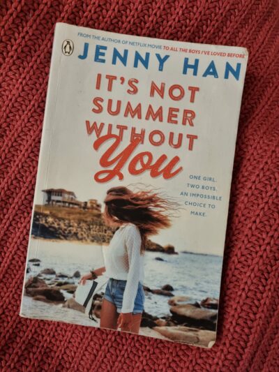 It's Not Summer Without You Cover. A girl on a beach in short denim shorts, with long hair, looking back towards a big beach house.