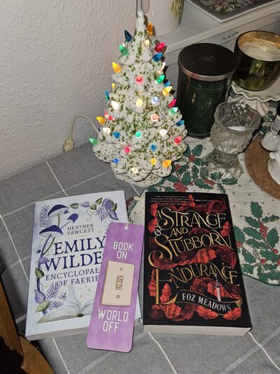 Picture of the two books Emily Wilde's Encyclopaedia of Faeries and A Strange and Stubborn Endurance sitting in front of a small porcelain Christmas tree with a bookmark