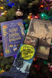 A picture of two books, "The Secret Lives of Country Gentlemen by KJ Charles and The Keeper's Six by Kate Elliot. Lying on the bottom of the books is a pair of socks that reads, "Dragons and Wizards and Shit".