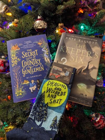 A picture of two books, "The Secret Lives of Country Gentlemen by KJ Charles and The Keeper's Six by Kate Elliot. Lying on the bottom of the books is a pair of socks that reads, "Dragons and Wizards and Shit".