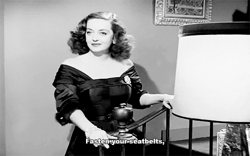 A gif from a classic Bette Davis movie, All About Eve. Bette Davis is standing on a staircase in a party dress. She says “Fasten your seatbelts.”
