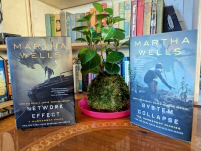 The books, "Network Effect" and "System Collapse" by Martha Wells standing on opposite sides of a green leafed plant growing out of a moss ball.