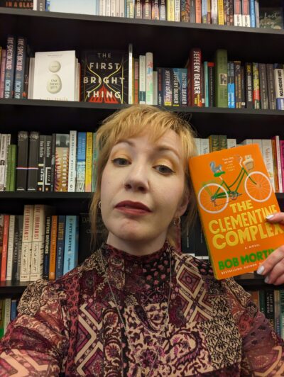 blonde woman in front of bookshelf holds a paperback copy of The Clementine Complex