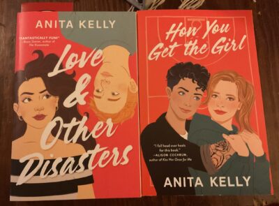 Anita Kelly’s books Love & Other Disasters and How You Get the Girl next to each other on a wooden table.