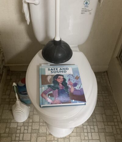 Picture of a white toilet, lid down. A plunger and a copy of Mercury Stardust’s Safe and Sound sit on the lid. This is the plunger I used on this toilet, following Stardust’s directions for a successful toilet unclogging. 
