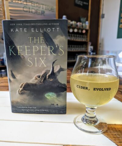 The book, "The Keeper's Six" by Kate Elliott standing next to a hard cider from Serpentine Cider.