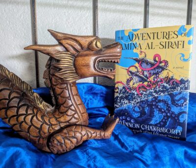 The book, "The Adventures of Amina al-Sirafi" by Shannon Chakraborty standing next to a wooden dragon-like sculpture, resting on shiny blue fabric giving the impression of water.