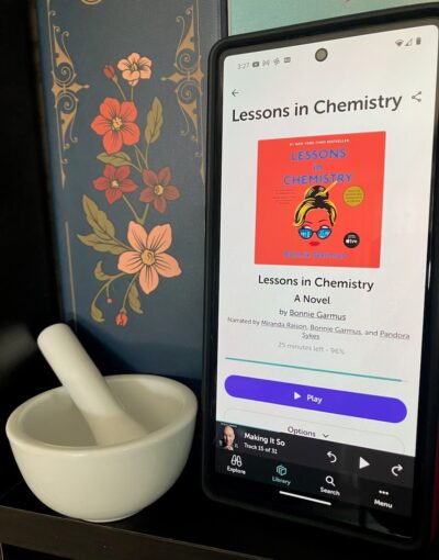 A cellphone showing the audiobook, "Lessons in Chemistry" next to a small mortar and pestle.