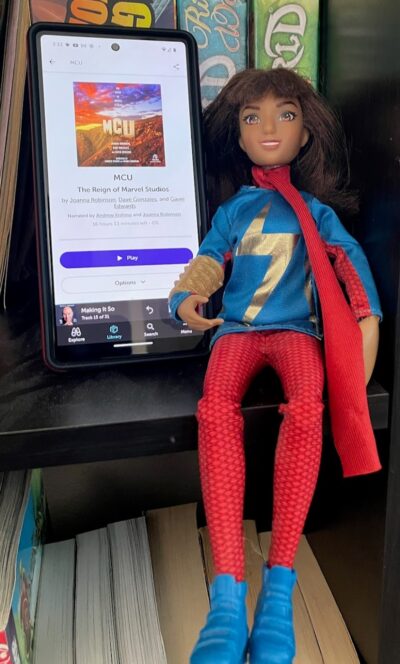 A cellphone showing the audiobook, "MCU: The Reign of Marvel Studios" next to a Ms. Marvel action figure.