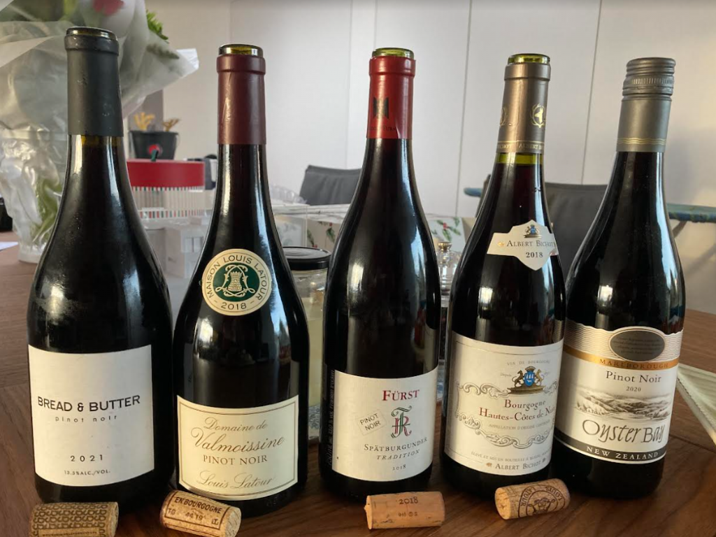 Five different pinot noir wines in bottles with their corks in front of them but one screw top still on