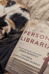A calico cat curled up sleeping next to the book, "The Personal Librarian" by Marie Benedict and Victoria Christopher Murray.