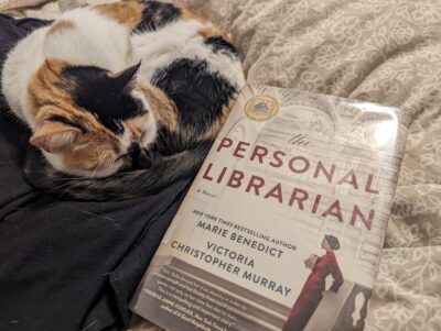 A calico cat curled up sleeping next to the book, "The Personal Librarian" by Marie Benedict and Victoria Christopher Murray.