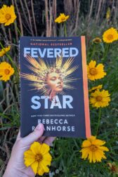 The book, "Fevered Star" by Rebecca Roanhorse held in front of yellow wildflowers.