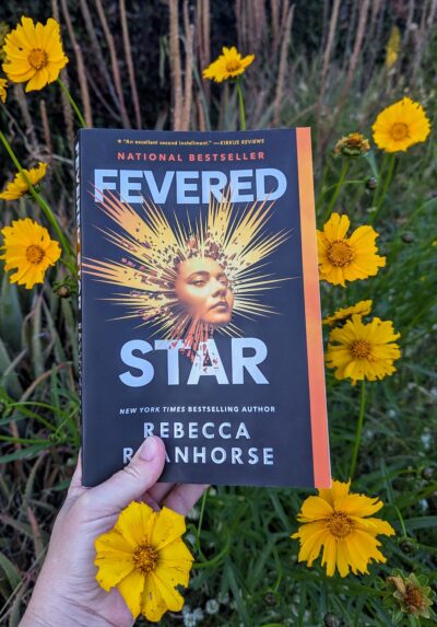The book, "Fevered Star" by Rebecca Roanhorse held in front of yellow wildflowers.