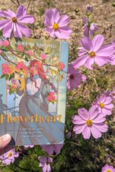 The book "Flowerheart" by Catherine Bakewell held in front of pale purple blooming cosmos flowers.