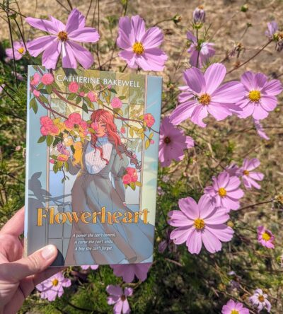 The book "Flowerheart" by Catherine Bakewell held in front of pale purple blooming cosmos flowers.