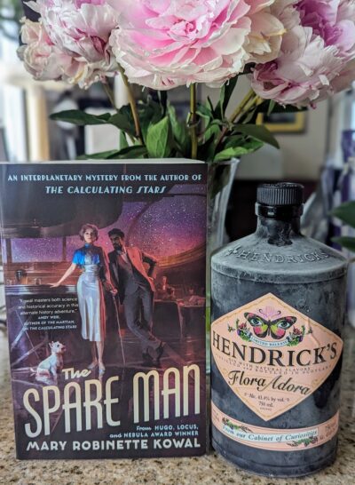 The book, "The Spare Man" by Mary Robinette Kowal standing next to a bottle of Hendricks, "Flora Adora" gin. A bunch of blooming pink peonies are in the background.