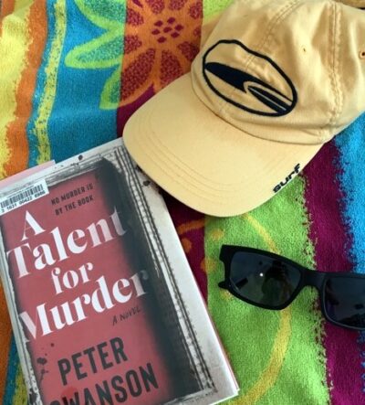 On a beach towel sit a cap, a pair of sunglasses, and a library book - Peter Swanson’s A Talent for Murder.