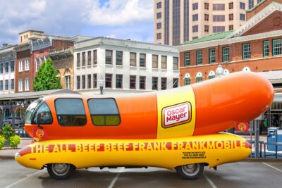 An Oscar Meyer wiener-mobile in a parking lot. The vehicle is shaped like a curved hot dog, but if it were painted beige, would look like a penis.