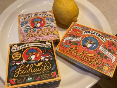 Three boxes of Fishwife Tinned Seafood Co. tinned fish sit on a plate with a lemon. The boxes are Albacore tuna in olive oil, slow smoked mackerel with chili flakes and smoked salmon with Szechuan chili crisp.