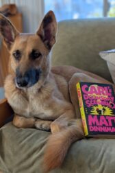 A German shepherd mix curled up in a chair with the book, "Carl's Doomsday Device" by Matt Dinniman resting on the dog's hindquarters.