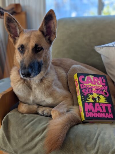 A German shepherd mix curled up in a chair with the book, "Carl's Doomsday Device" by Matt Dinniman resting on the dog's hindquarters.