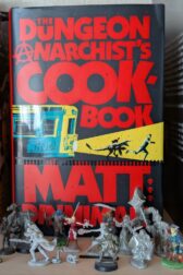 The book, "The Dungeon Anarchist's Cookbook" by Matt Dinniman is standing behind an assortment of miniature figures used in roleplaying games like Dungeons and Dragons.