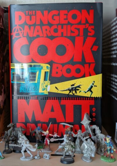 The book, "The Dungeon Anarchist's Cookbook" by Matt Dinniman is standing behind an assortment of miniature figures used in roleplaying games like Dungeons and Dragons.