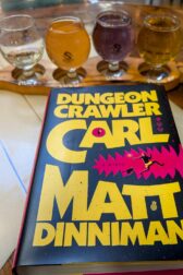 The book, "Dungeon Crawler Carl" lying down in front of a flight of 4 small cider glasses full of colorful cider.