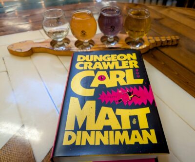 The book, "Dungeon Crawler Carl" lying down in front of a flight of 4 small cider glasses full of colorful cider.