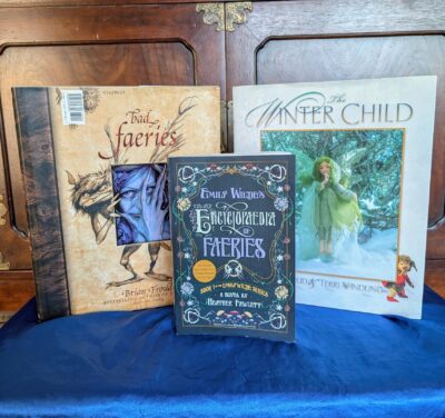 The book, "Emily Wilde's Encyclopedia of Faeries" by Heather Fawcett is standing in front of two books with fairies on their covers.
