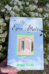 The book, "The Eyre Affair" by Jasper Fforde is being held up in front of a patch of small white alyssum flowers.