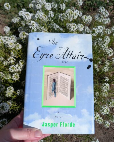 The book, "The Eyre Affair" by Jasper Fforde is being held up in front of a patch of small white alyssum flowers.
