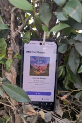 A phone with the Libro Fm reading app open to "Into the Woods" by Jenny Holiday. The phone is tucked into a hedge with green leaves.