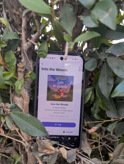 A phone with the Libro Fm reading app open to "Into the Woods" by Jenny Holiday. The phone is tucked into a hedge with green leaves.