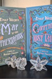 The books Emily Wilde's Map of the Otherlands and Emily Wilde's Compendium of Lost Tales written by Heather Fawcett are standing on a bookshelf. In front of them are a small rose and daffodil made out of crystal.