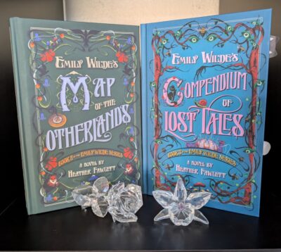 The books Emily Wilde's Map of the Otherlands and Emily Wilde's Compendium of Lost Tales written by Heather Fawcett are standing on a bookshelf. In front of them are a small rose and daffodil made out of crystal.