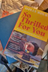 Cover of So Thrilled For You, yellow and pink, shows a woman lying on a floor wearing a party hat blowing out a candle on a cupcake