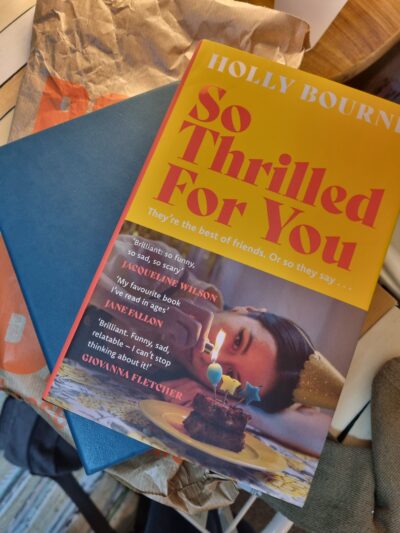 Cover of So Thrilled For You, yellow and pink, shows a woman lying on a floor wearing a party hat blowing out a candle on a cupcake