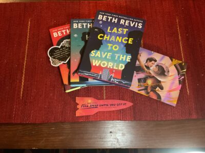 On a red woven table runner, Beth Revis’s Last Chance to Save the World sits on a fanned stack of the Chaotic Orbit series books. On the left is a sticker in the shape of a valentine with text reading “roses are red. Outerspace is black. Billionaires shouldn’t exist. Capitalism is whack.”