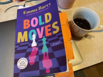 A physical advance reader copy of Emma Barry’s Bold Moves sits on top of a laptop, with orange tabs bristling from the side.