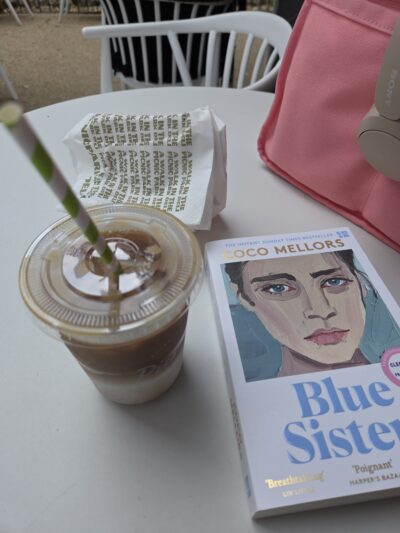 Copy of Blue Sisters, which has the painted face of a woman, on a table with an iced coffee 