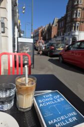 street scene with copy of Song of Achilles on a table with an iced coffee