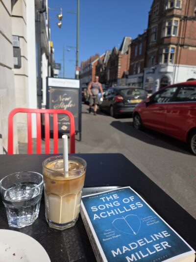 street scene with copy of Song of Achilles on a table with an iced coffee
