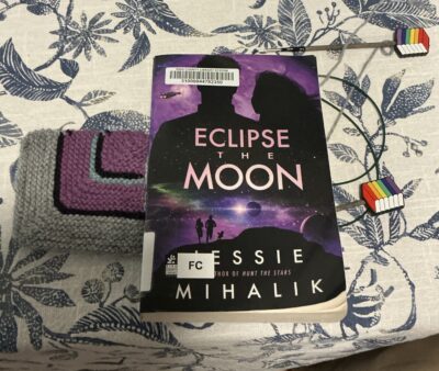 knitted mitered square with grey border and stripes of black, purple, and teal next to a paperback book with the colors purple, black, and teal on the cover. Both sitting on a table with a white and blue tablecloth.